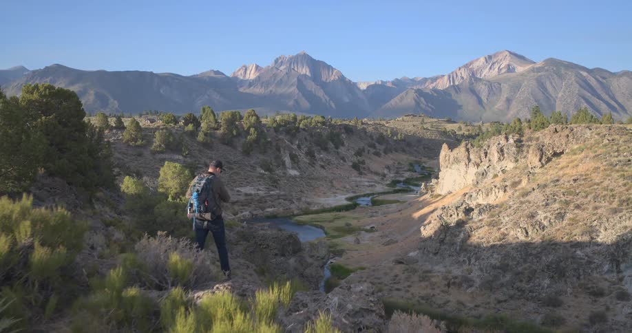 A hiker walks along a cliff's edge revealing a scenic river valley and a majestic mountain range in the distance