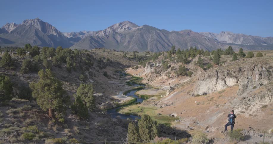 A photographer with a backpack hikes to a viewpoint to capture the stunning landscape of Hot Creek Gorge with the Sierra Nevada mountains in the background