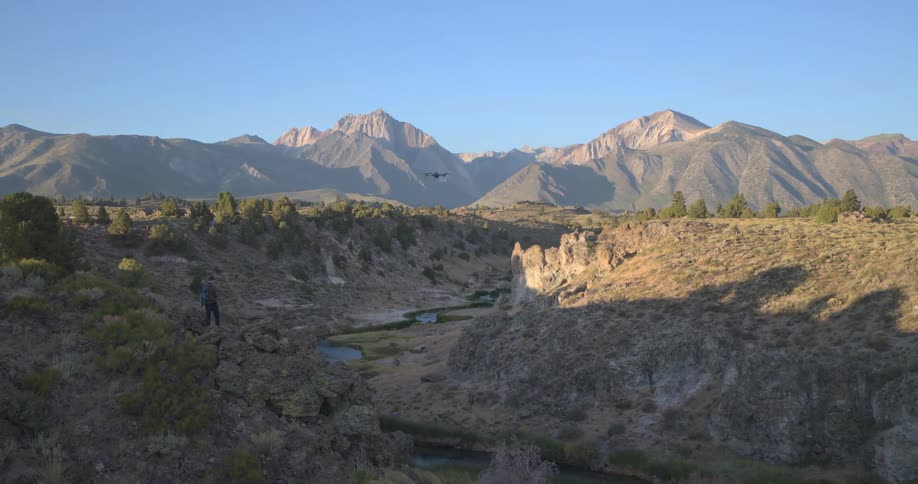 A hiker operates a drone, flying it over a river canyon against a backdrop of majestic mountains at sunrise