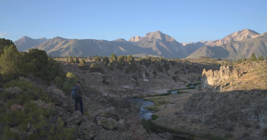 A man with a backpack watches his drone fly over a beautiful mountain river valley during golden hour