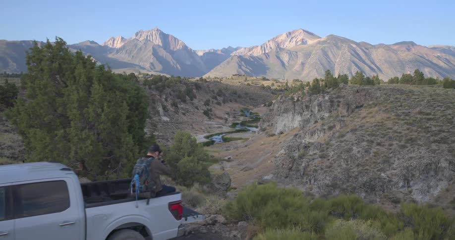 An adventurer sits on his pickup truck enjoying the mountain view before a drone reveals a stunning river canyon