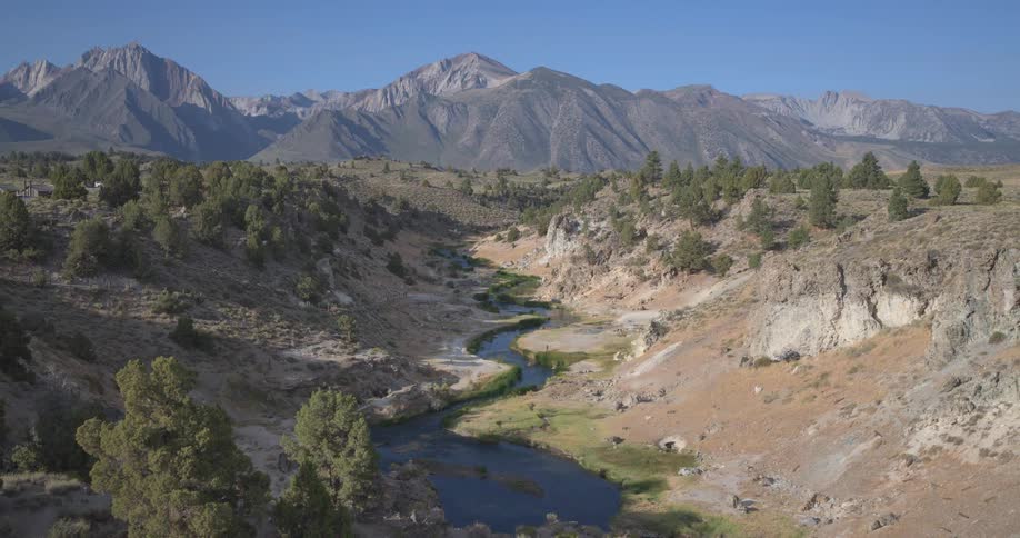 A lone hiker traverses a rugged trail overlooking a winding river in a vast mountain valley under a clear blue sky