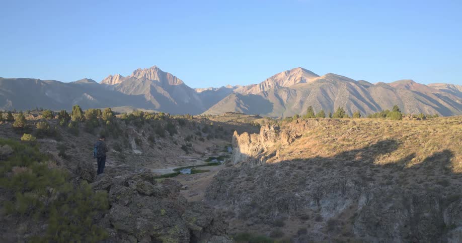 A hiker stands on a rocky cliff overlooking a canyon and flies a drone to capture the scenic mountain landscape
