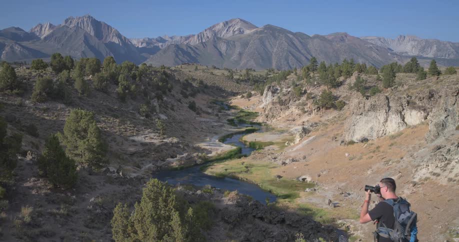 A male photographer with a backpack captures the stunning view of a winding river in a valley with majestic mountains in the background
