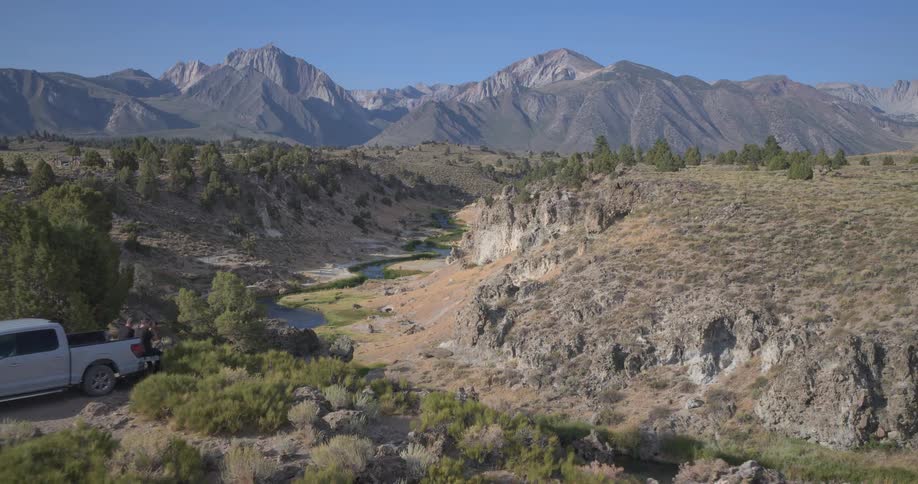 A silver pickup truck is parked next to a winding river in a rugged canyon with a majestic mountain range in the background