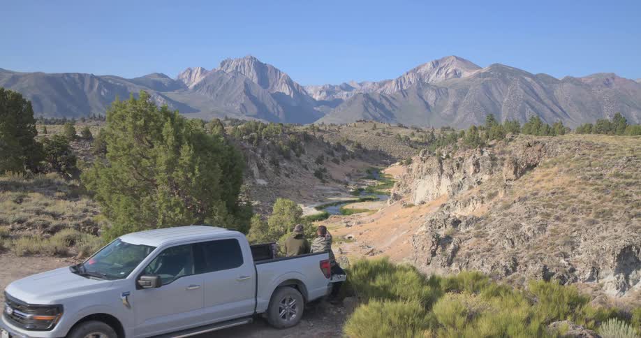 An aerial view slowly pushes in on a couple relaxing on the tailgate of their truck, enjoying the breathtaking scenery of a mountain valley