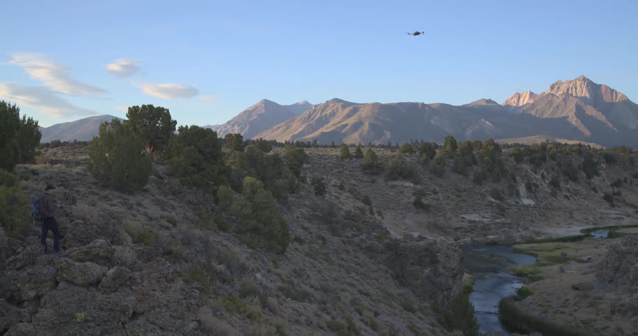 A man flies a drone in slow motion over a river in a majestic mountain valley during a golden sunset