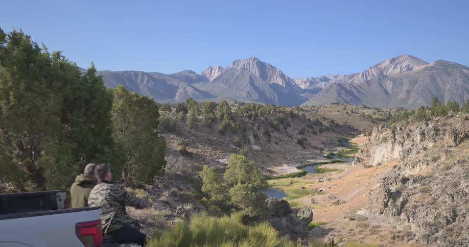 Slow zoom past two men sitting on their truck's tailgate as they enjoy the majestic mountain and river valley view