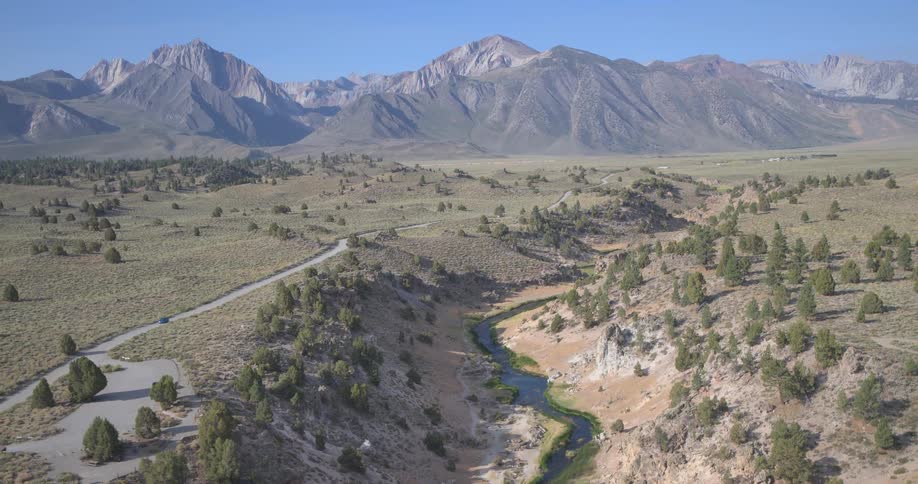 A sweeping aerial view pulls back to reveal a river winding through a vast valley with geothermal hot springs at the foot of the Sierra Nevada mountains