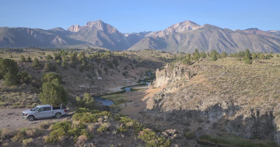 Aerial view pulls back to show two people relaxing on the tailgate of a truck, enjoying the majestic mountain and river valley scenery