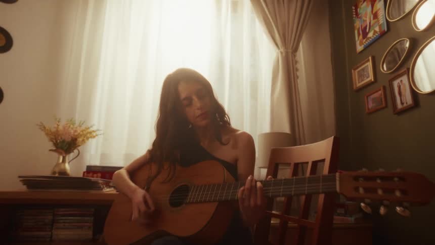 A young woman sits in her cozy, sunlit room and soulfully plays the acoustic guitar
