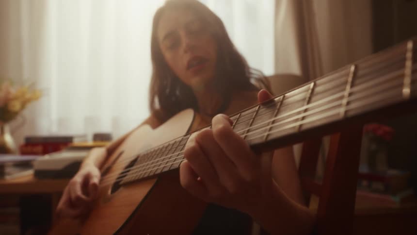 A young woman passionately sings and plays an acoustic guitar in her sun-drenched room, lost in the music