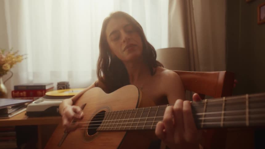 A young woman plays an acoustic guitar and sings softly in her warmly lit room