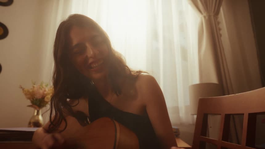 A joyful woman with long dark hair laughs contagiously while playing an acoustic guitar in a warmly lit room