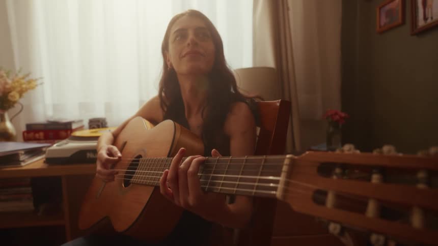 A young woman with long dark hair sits in her warmly lit room and practices playing a classical guitar by the window