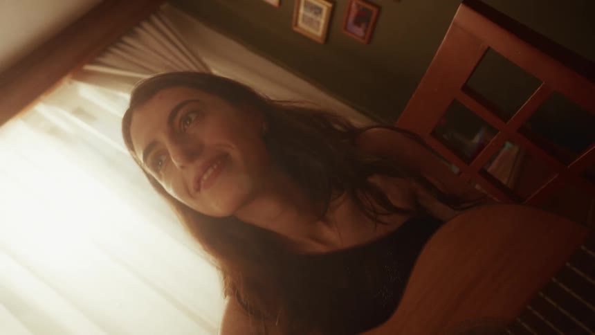 A talented female musician passionately plays her acoustic guitar and sings while sitting in her cozy, sun-drenched room