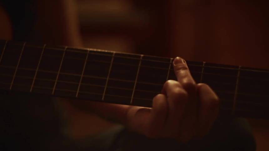 Close-up on a guitarist's hand fretting chords on a classical guitar in a dimly lit, warm environment