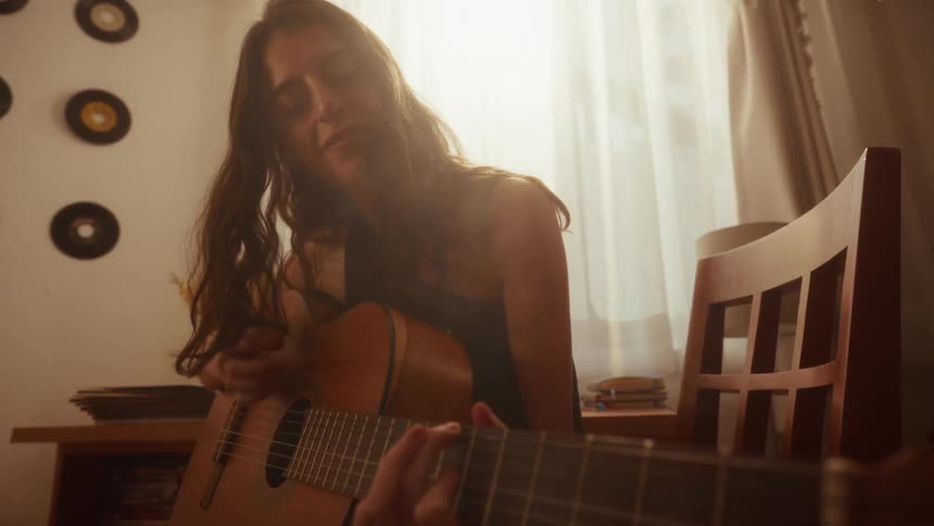A soulful young woman plays her acoustic guitar, bathed in the warm, hazy light from a window in her cozy room