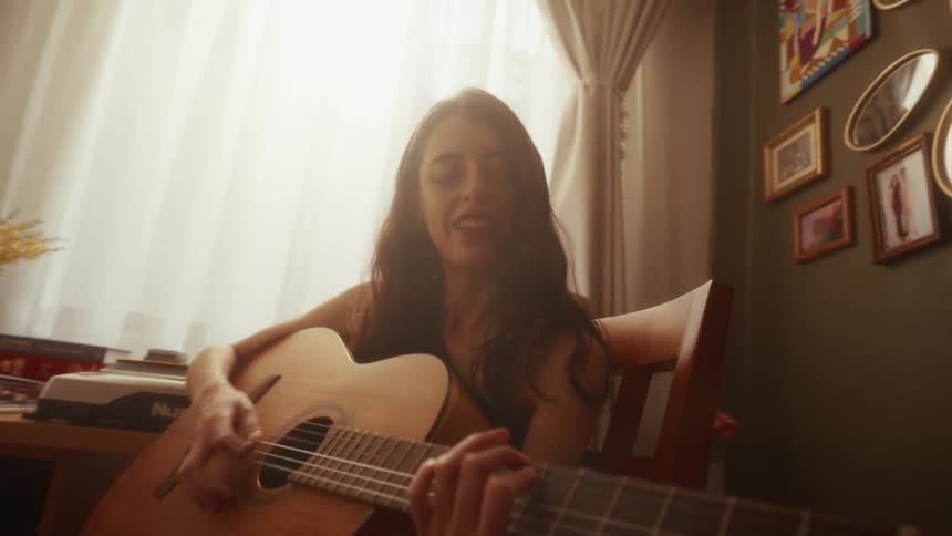 A smiling young woman sits in a sunlit room, joyfully playing an acoustic guitar and singing