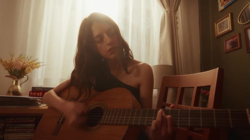 A woman with her eyes closed passionately plays a heartfelt song on an acoustic guitar in a cozy, softly lit room