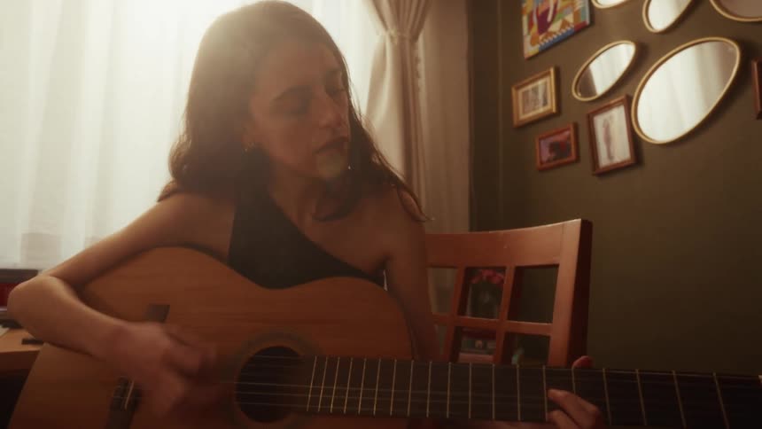 A woman passionately sings and plays the acoustic guitar in her cozy, sunlit room