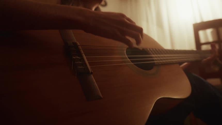 Close-up shot of a musician's hands strumming an acoustic guitar in a warmly lit, cozy room