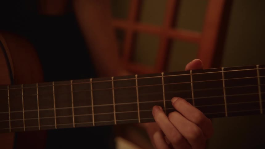 A close-up of a musician's hand playing chords on the fretboard of an acoustic guitar in a warmly lit room