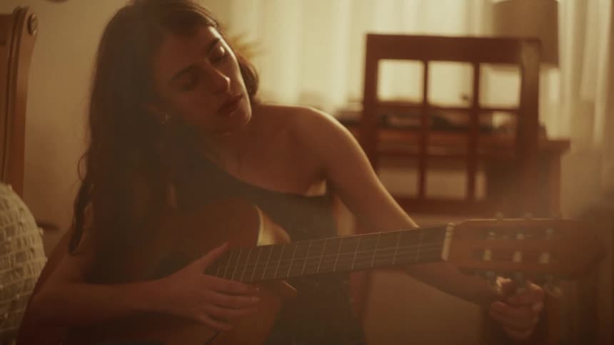 A focused young woman with long dark hair tunes her acoustic guitar in a warmly lit, hazy room