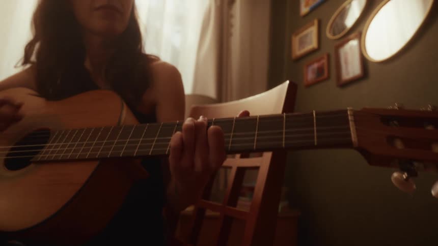 A focused young woman plays an acoustic guitar in her cozy, sun-drenched room, lost in the melody