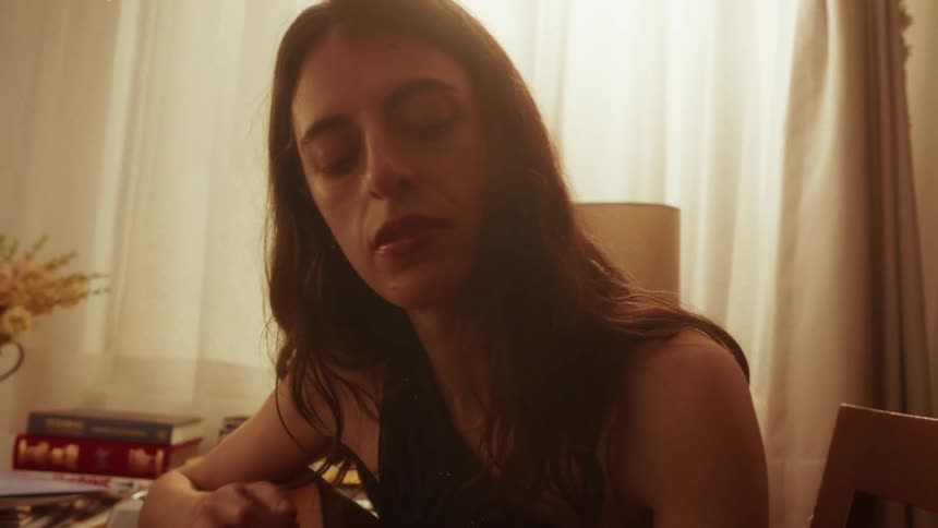 A contemplative woman with her eyes closed plays an acoustic guitar in a warmly lit room in slow motion