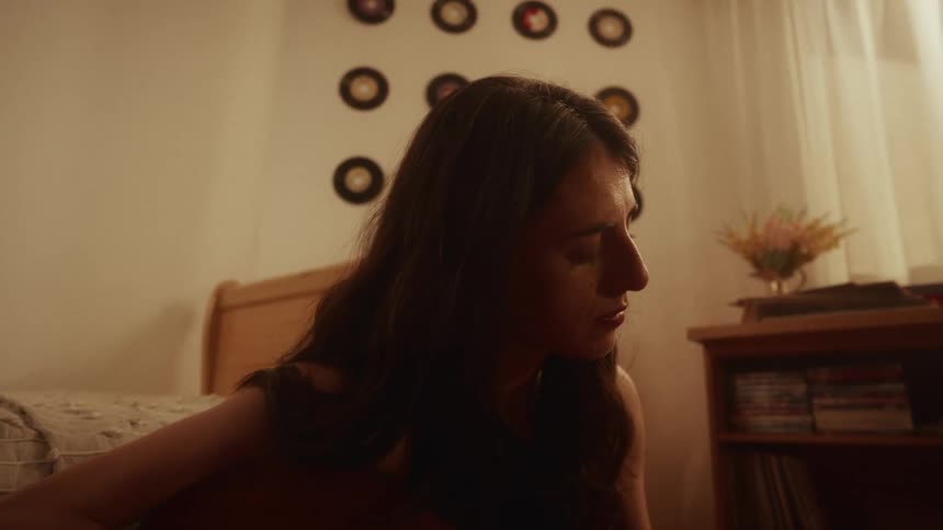 A slow push-in shot reveals a young woman playing an acoustic guitar in her warmly lit bedroom, surrounded by her collection of classic vinyl records