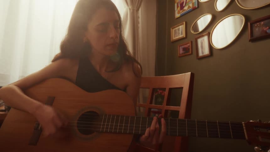 A serene woman with long dark hair strums an acoustic guitar by a window, bathed in warm afternoon light