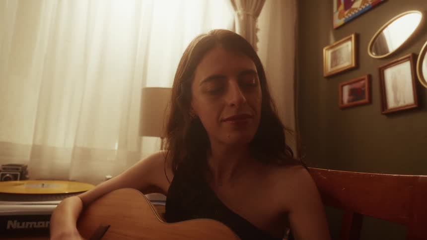 A young woman sits in her warmly lit room and peacefully plays an intimate melody on her acoustic guitar