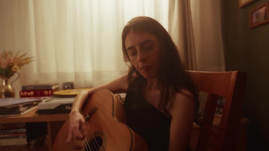 A woman sits in a warmly lit room and pensively plays a slow melody on her acoustic guitar