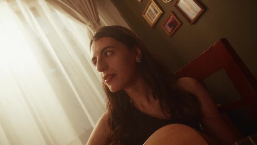 The camera pushes in on a woman playing an acoustic guitar in her warmly lit room, she stops playing and gazes out the window