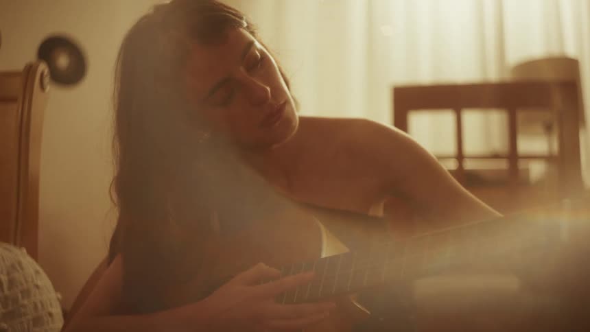 A serene woman with long dark hair peacefully plays an acoustic guitar in a warmly lit room