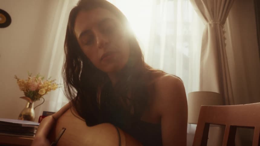 A female musician serenely plays her acoustic guitar in a sun-drenched room, with light softly filtering through the window behind her