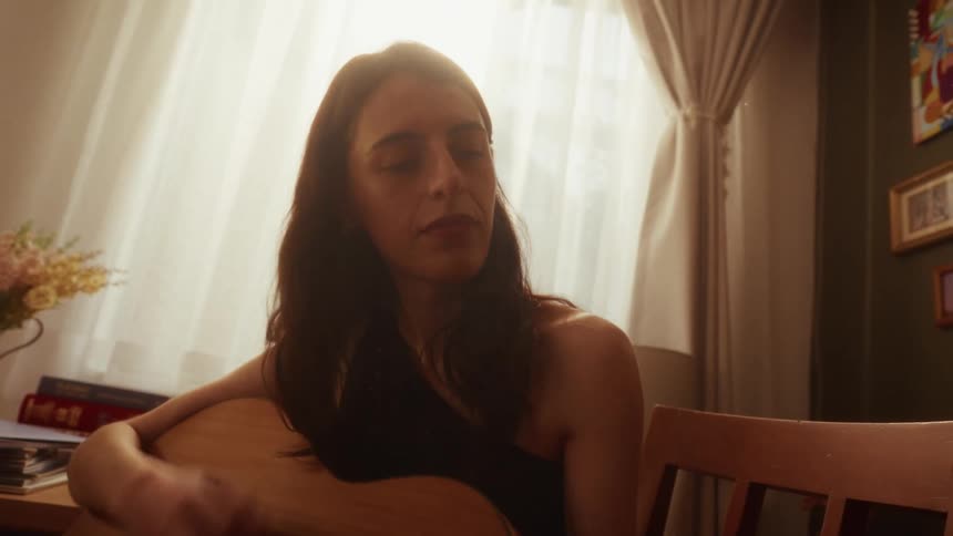 A thoughtful woman plays a melancholic tune on her acoustic guitar in a sun-drenched room, her face illuminated by the warm light from the window
