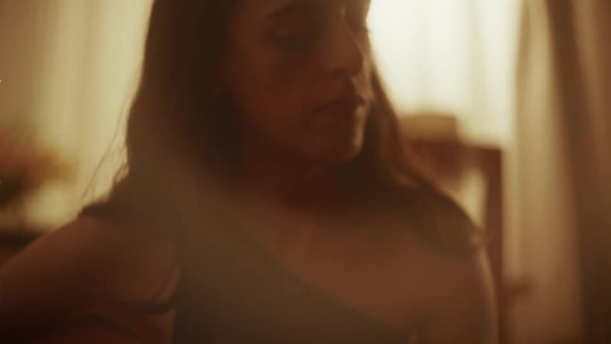 A woman with her eyes closed plays an acoustic guitar in a softly lit, hazy room
