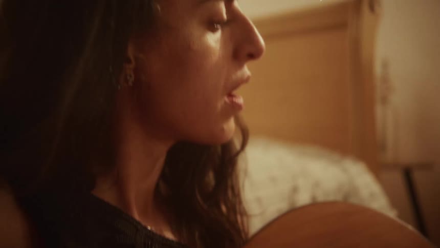 An intimate slow-motion close-up of a woman singing and strumming an acoustic guitar in her bedroom