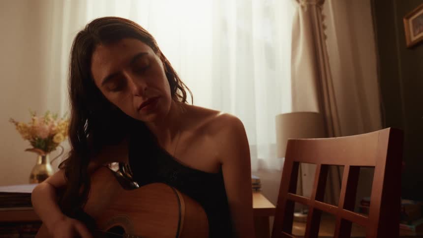 A woman with her eyes closed passionately plays a melancholic melody on her acoustic guitar in a warmly lit room
