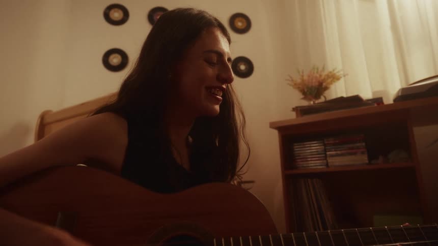 A joyful young woman with long dark hair smiles as she sings and plays an acoustic guitar in her warmly lit room
