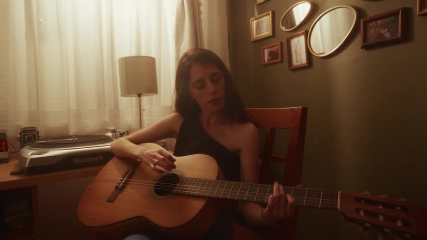 A woman plays an acoustic guitar with concentration while sitting in a warmly lit, cozy room