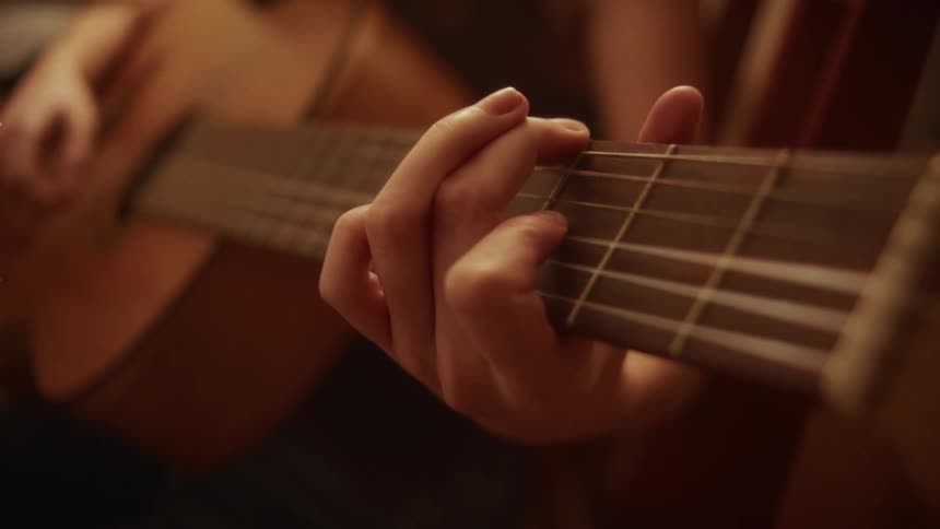 A musician's hands play chords on an acoustic guitar, with a rack focus bringing the fretboard into sharp detail