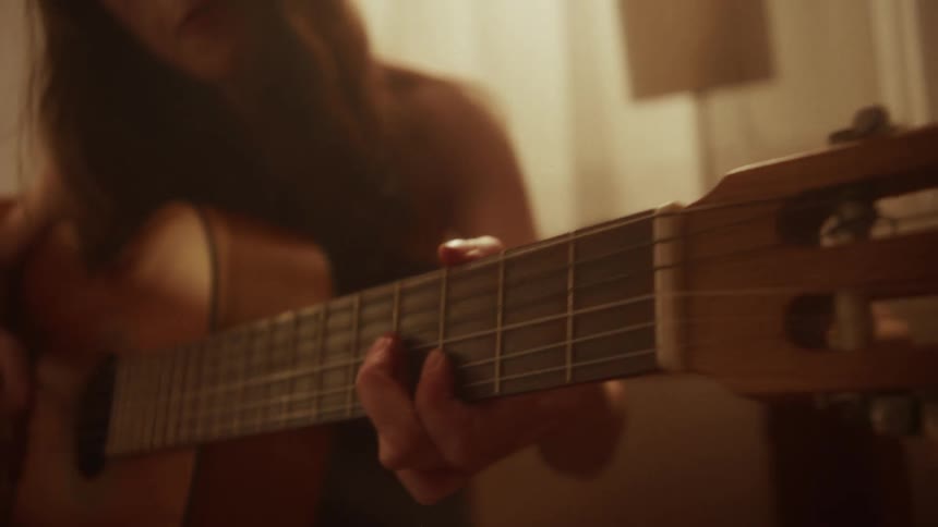 An intimate close-up shot pans along an acoustic guitar being played by a woman in a warmly lit room