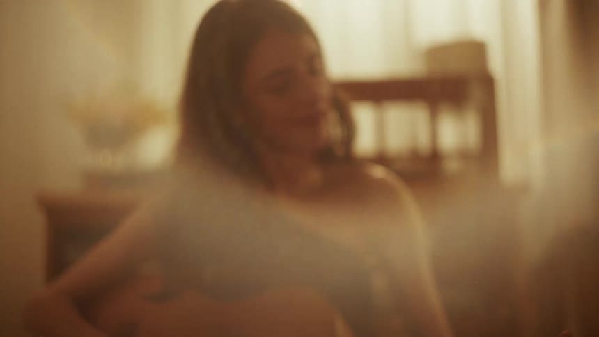 A young woman sits cross-legged on the floor surrounded by vinyl records, playing an acoustic guitar in a hazy, warmly lit room