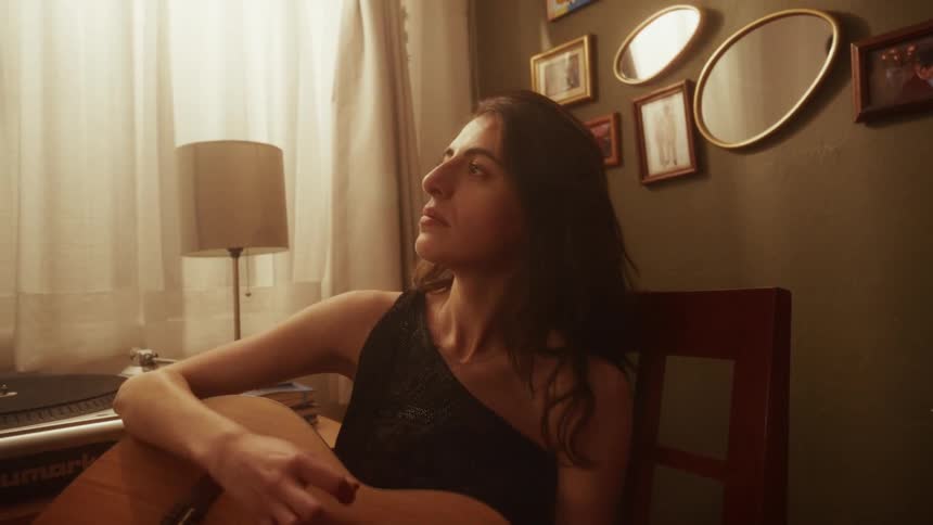 A beautiful woman thoughtfully plays an acoustic guitar in a warmly lit room with an emotional expression