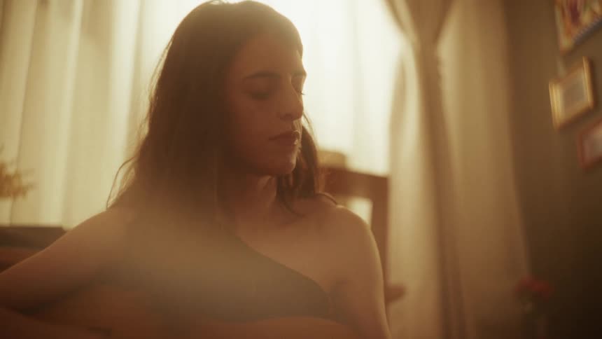 A young woman soulfully plays her acoustic guitar with her eyes closed in a warmly lit, intimate room