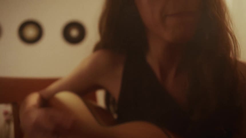 An artistic, soft-focus shot pans from a woman's hands strumming a guitar to her face, bathed in warm light