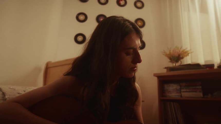 A young woman sits on the floor of her cozy bedroom, surrounded by vinyl records, as she plays the acoustic guitar and sings